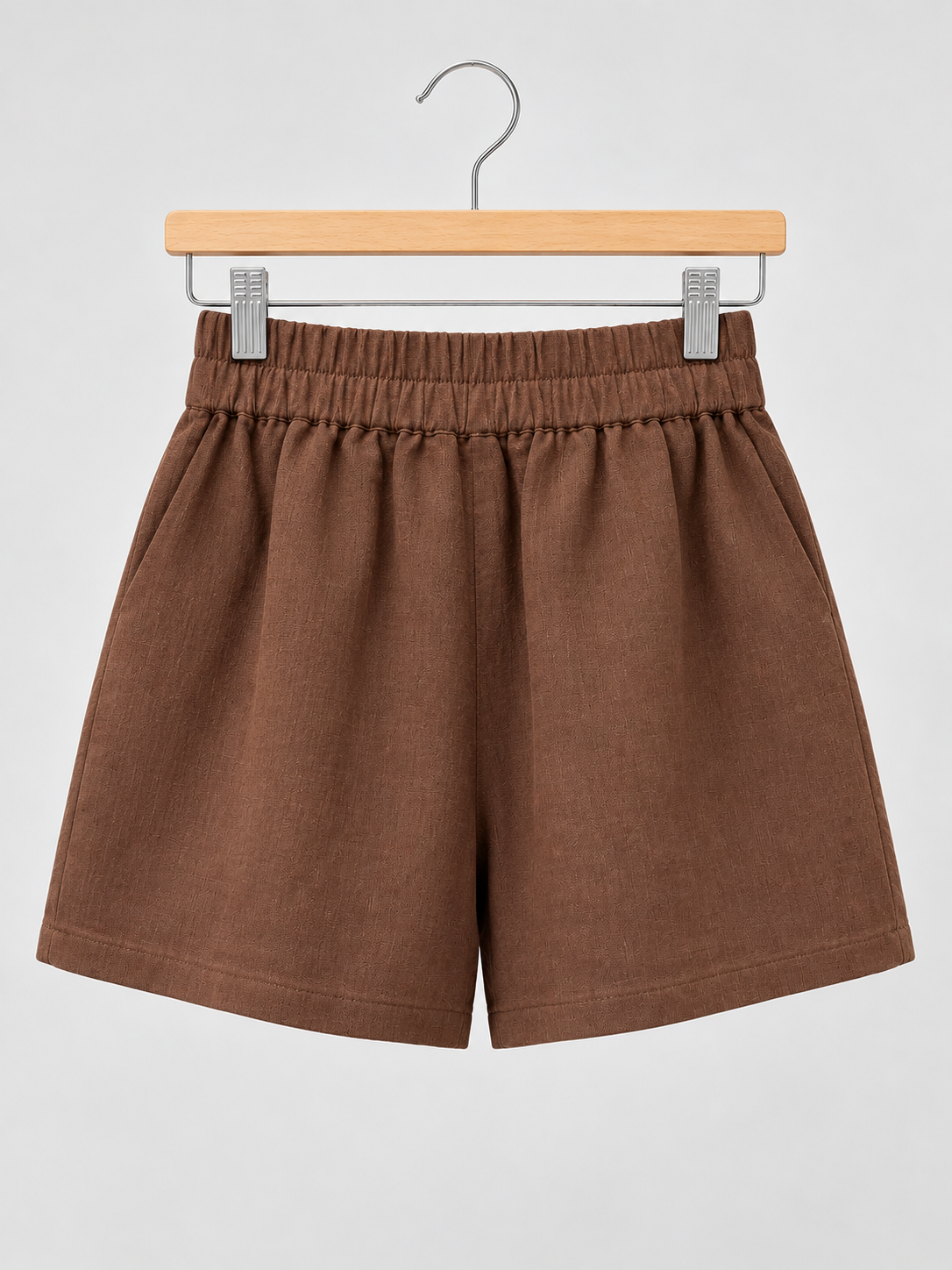Brown linen-blend shorts with an elastic waistband and side pockets, displayed on a wooden hanger against a light grey background.